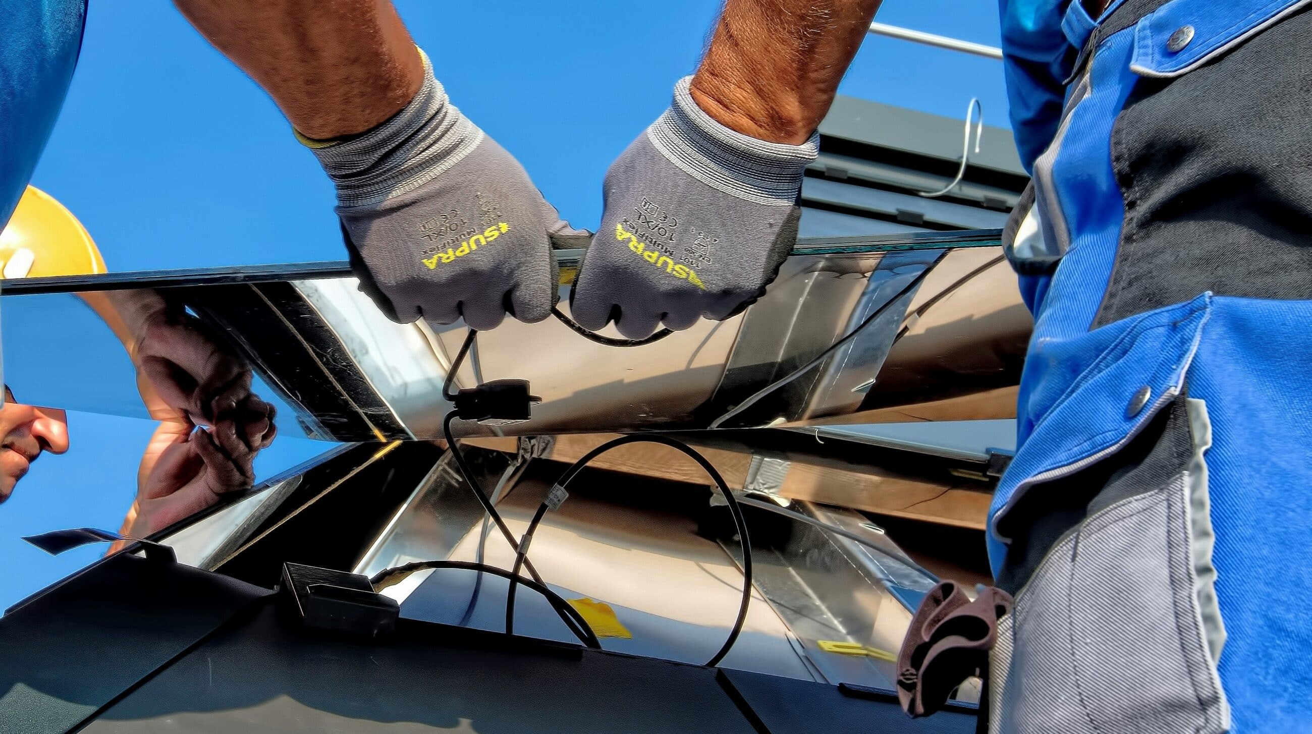 Handwerker montiert Photovoltaikmodule auf einem Dach, Nahaufnahme der Hände mit Schutzhandschuhen beim Anschließen der Solarkabel